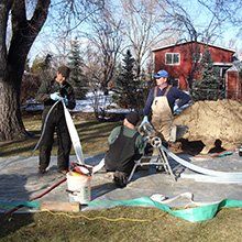 Three workers installing a drain system in a yard. One feeds a white tube, another kneels, and a third looks on. Sunny day.