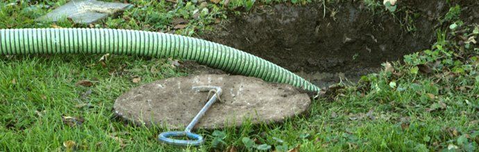A green hose lies on grass near an open hole, a manhole cover, and a lid-lifting tool.