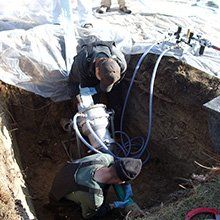 Two workers in a trench inspecting a metal component with tubes connected. Plastic sheet covers the ground.