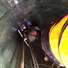 Person inside a large, dark-colored pipe, possibly a sewer. They look up, a yellow safety vest is visible.
