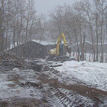 Yellow excavator digging in snow-covered construction site, trees in background, during snowfall.