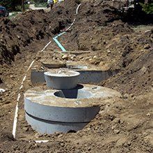 Excavated ground with concrete septic tank components and piping laid in trench.
