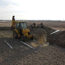 Yellow backhoe digging in a field, moving dirt near white pipes.
