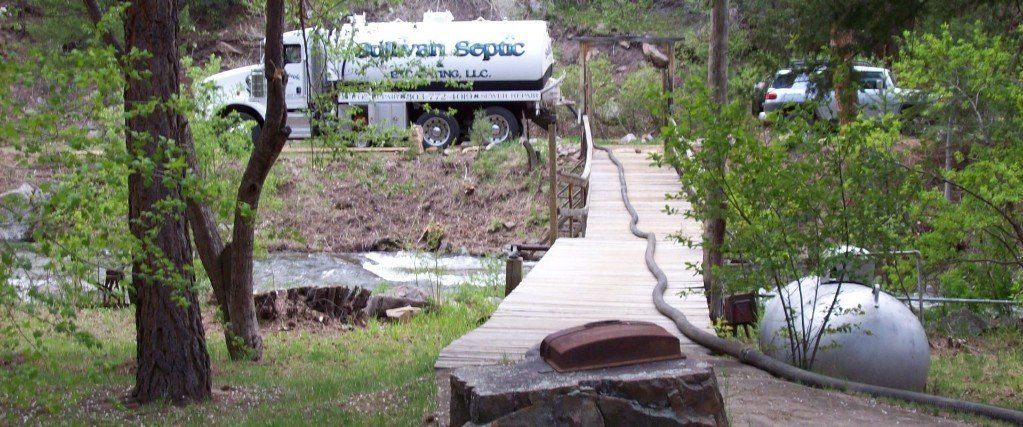 A septic truck parked near a bridge with a propane tank, surrounded by trees.
