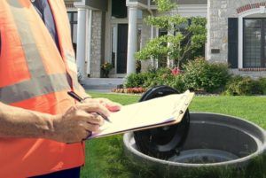 Person in orange vest taking notes near an open septic tank in front of a house.