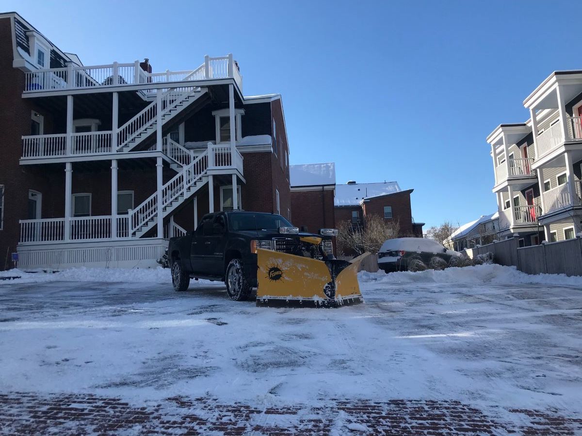 A snow plow is plowing a parking lot in front of a building.