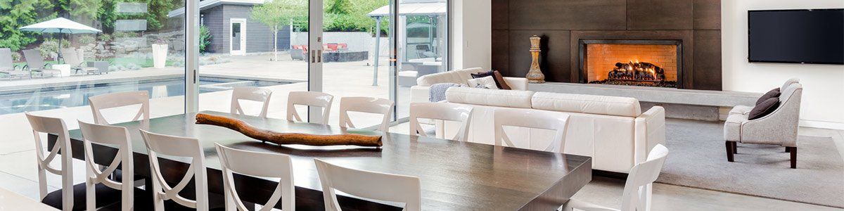 Modern living room with white sectional sofas, glass table, and a fireplace overlooking the outdoors