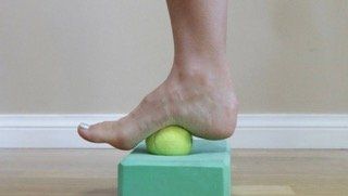 A person is standing on a yoga block with their foot on a tennis ball.