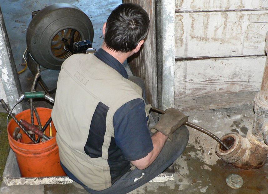 The image shows a plumber using a plumber's snake, also known as a drain auger, to clear a clogged pipe.