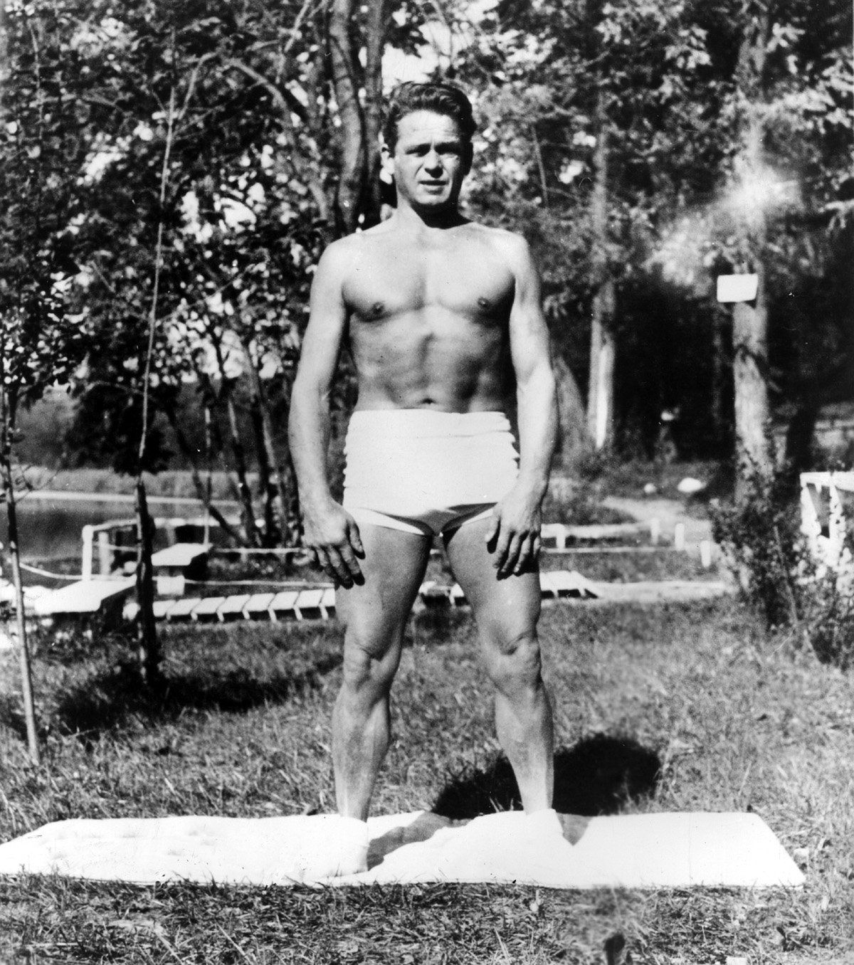 Joe Pilates standing without a shirt is standing on a Pilates mat