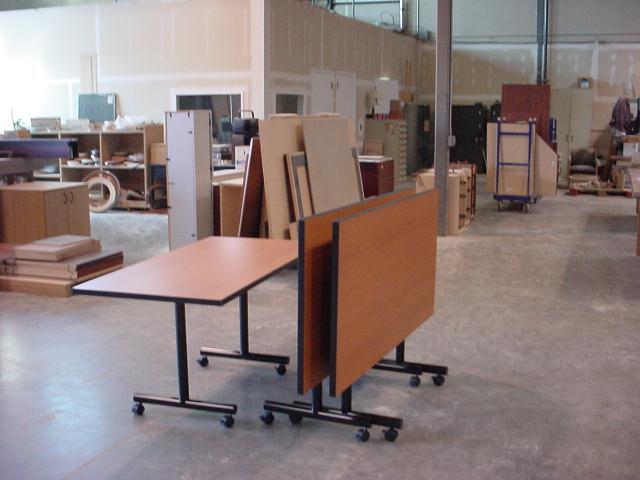 Two brown folding tables with black legs and wheels, in a cluttered workshop setting.