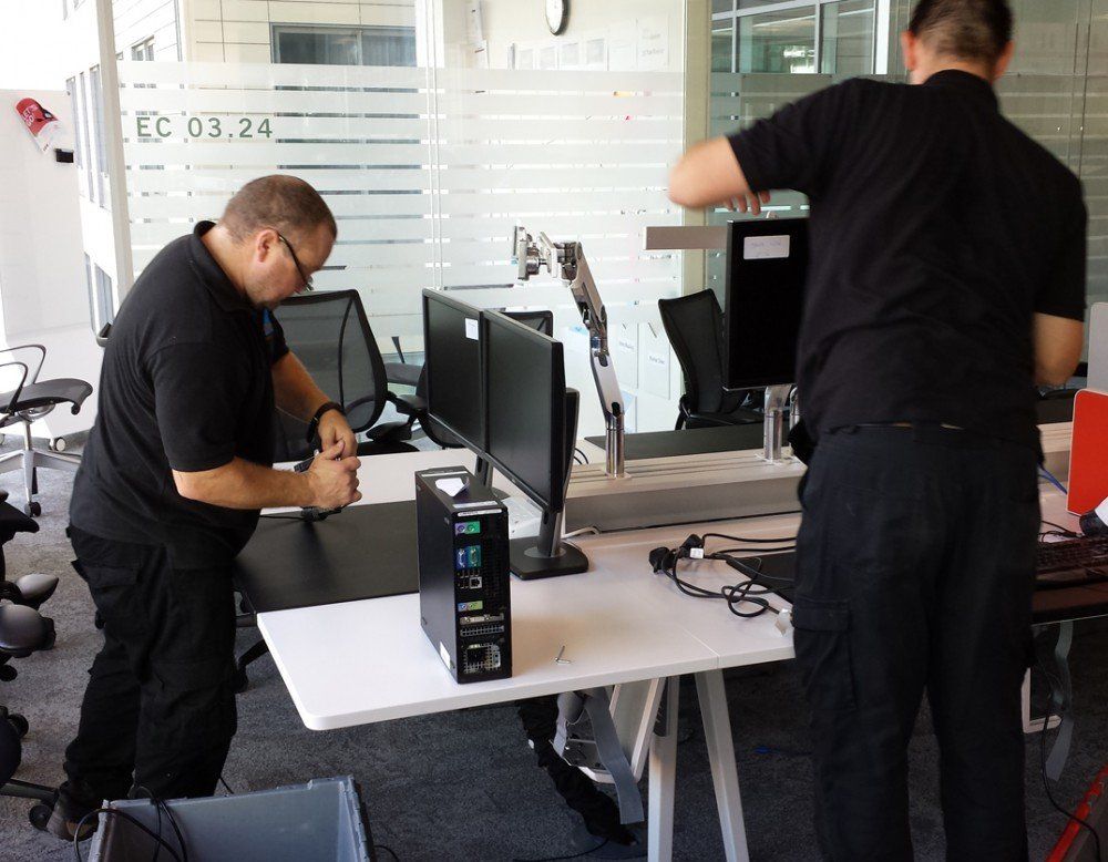 Two men are working on a computer in an office with a sign that says ec on it