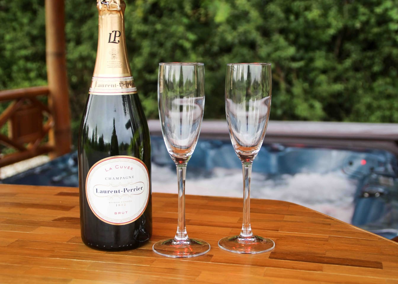 A bottle of champagne and two glasses are on a wooden table in front of a hot tub.