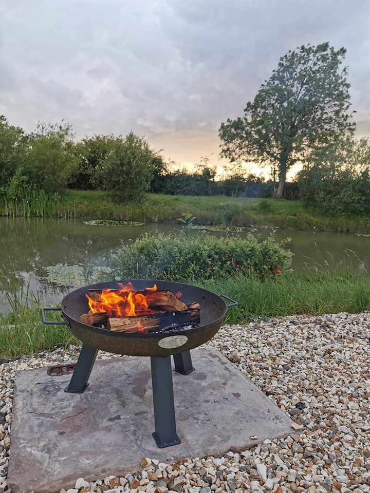 A fire pit is sitting next to a pond with a tree in the background.