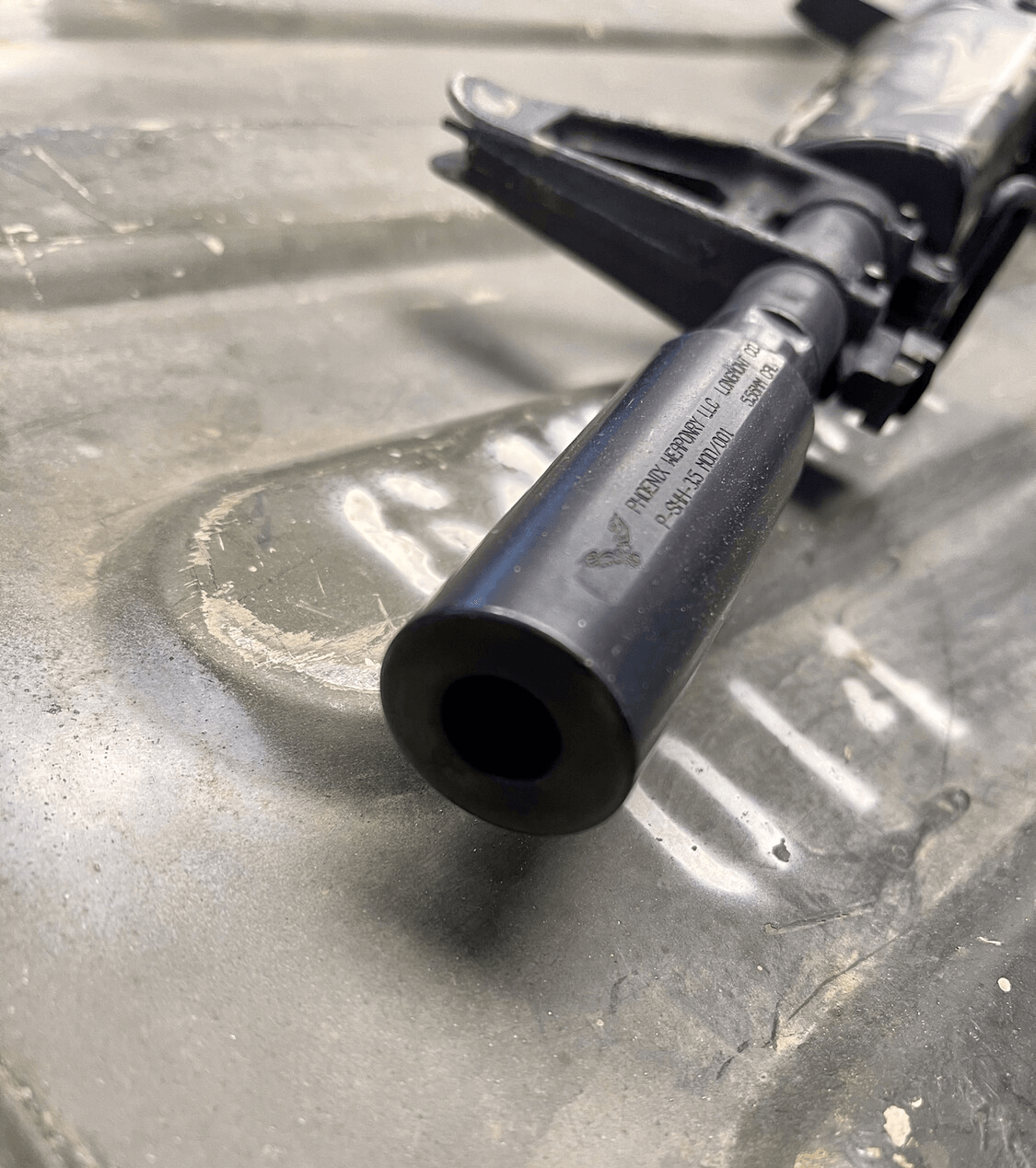 Close-up of a black rifle muzzle with visible text; resting on an olive drab surface.