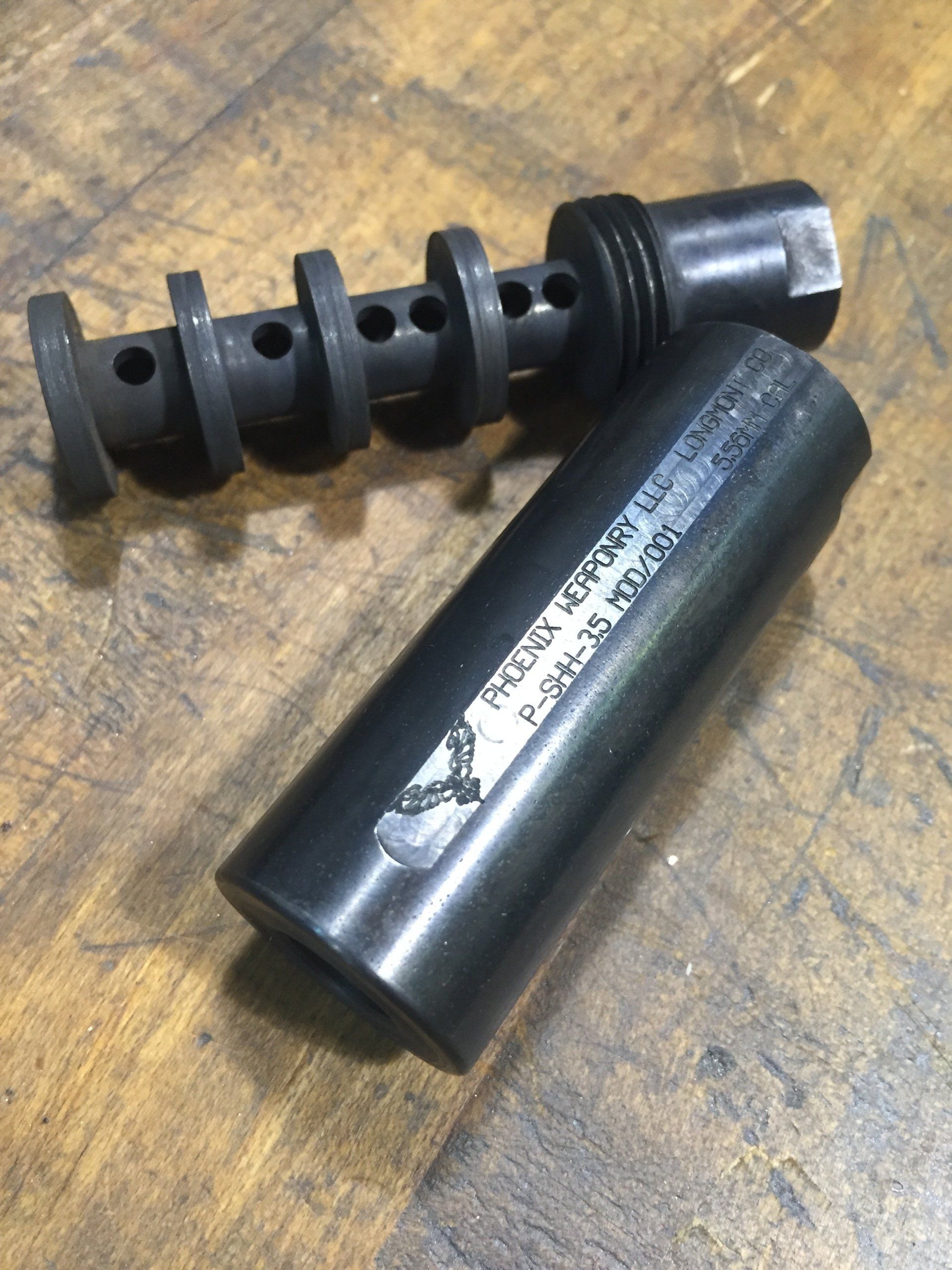 Two-piece, dark gray firearm suppressor. One piece is a ported barrel, the other a cylindrical sleeve with stamped markings.