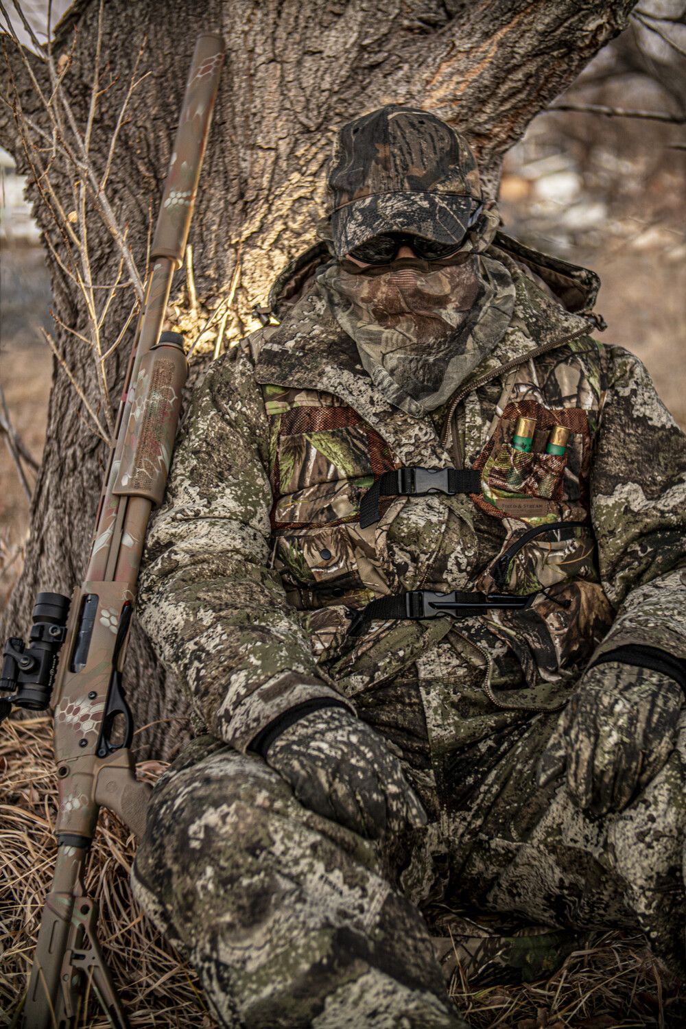 Hunter in camouflage sits leaning against a tree, shotgun at side.