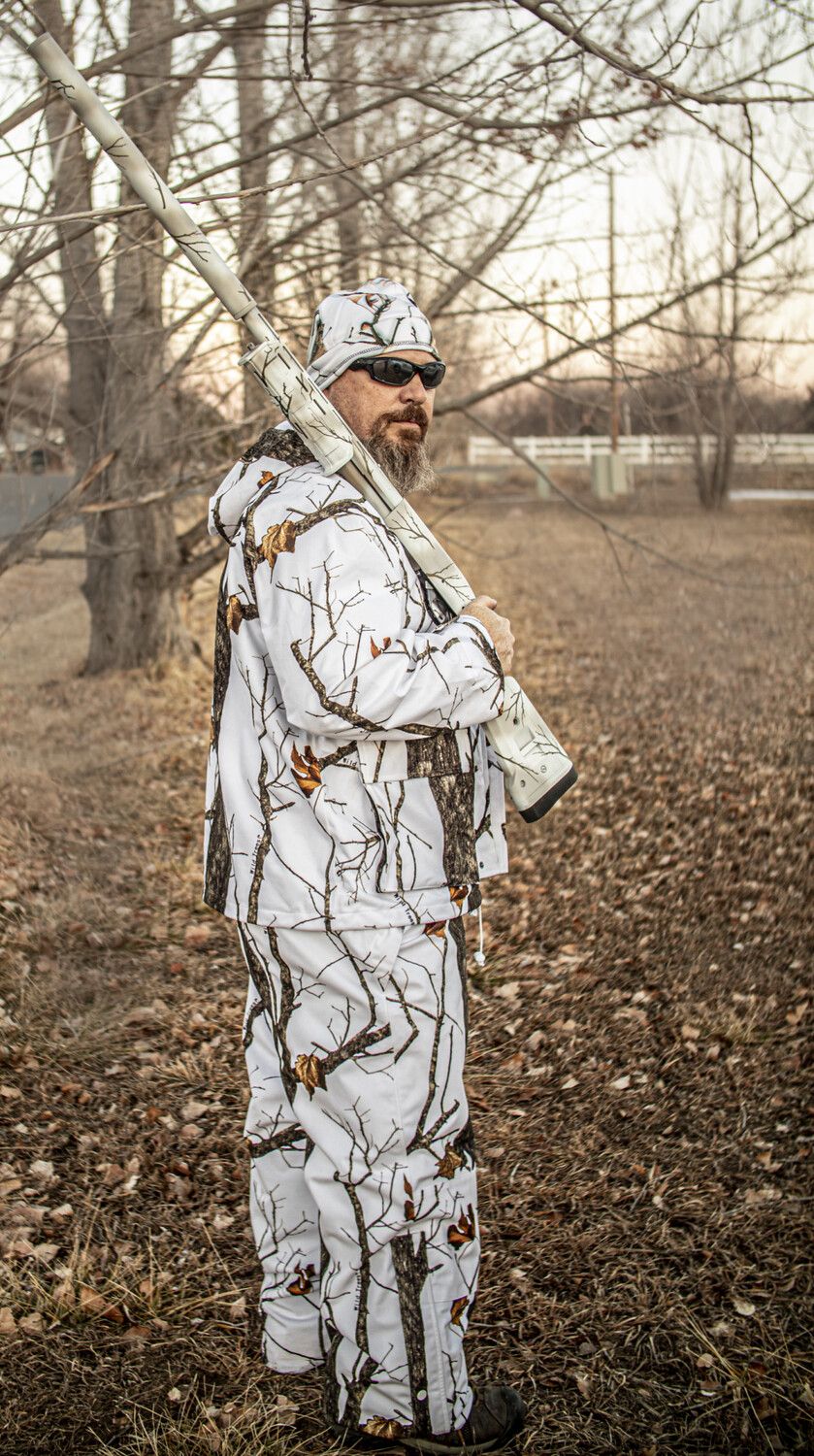 Man in white camouflage holding a shotgun outdoors near bare trees and dry leaves.