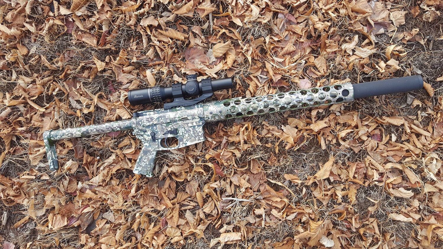 Integrally Suppressed AR-15 Platform Rifle Morgan Camouflaged rifle with scope and suppressor on fallen leaves.