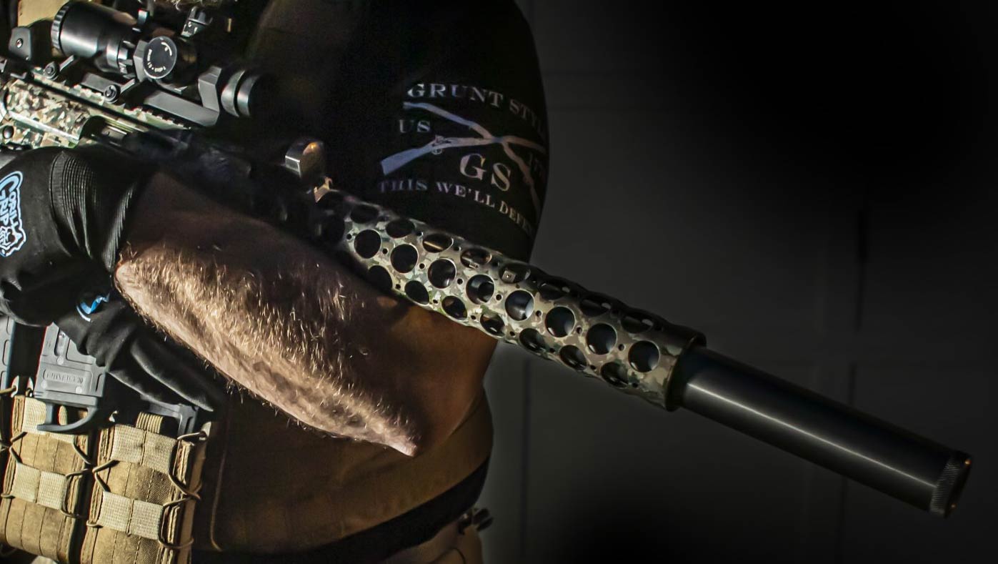 Person holding a rifle with a patterned barrel and scope, silencer attached.