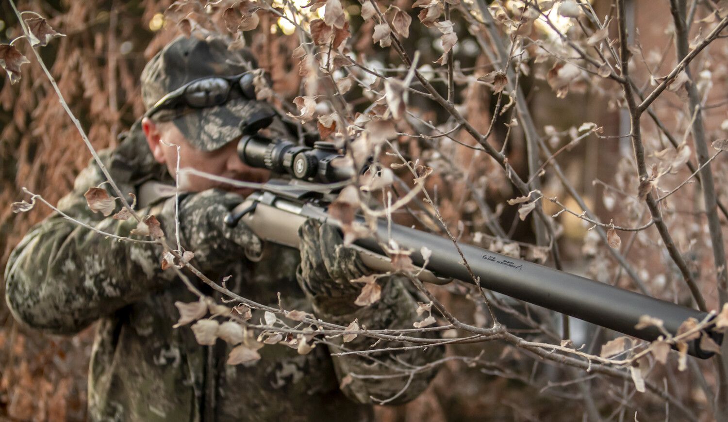 Hunter aiming rifle through a scope, camouflaged in a wooded area.