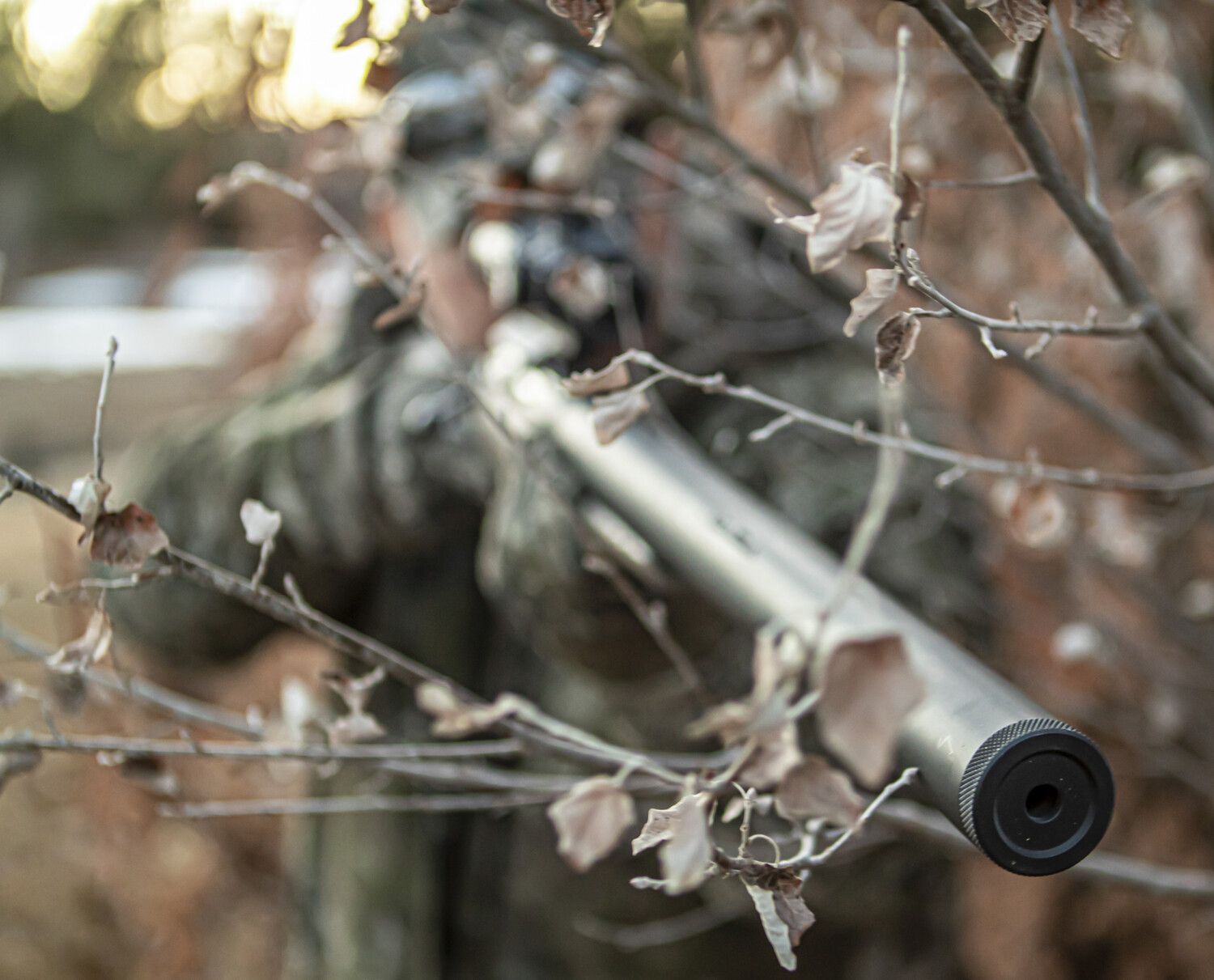 Hunter in camouflage aims a rifle from behind branches, outdoors.