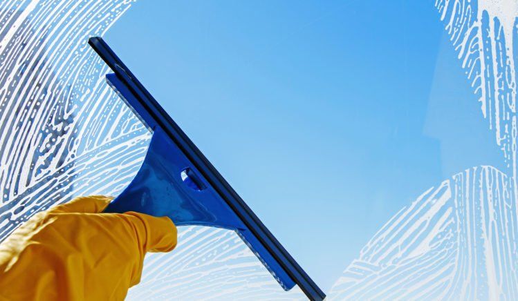 Hand in yellow glove uses a blue squeegee to clean a soapy window, revealing a clear blue sky.