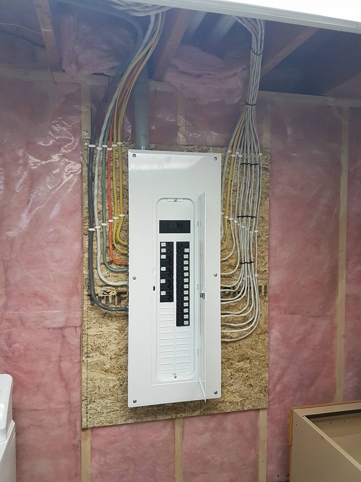 A wall-mounted electrical breaker panel centered on a wooden board, surrounded by pink fiberglass insulation.