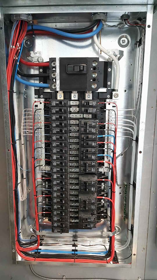 An open electrical service panel showing numerous black circuit breakers, red and blue wiring, and grounding connections.
