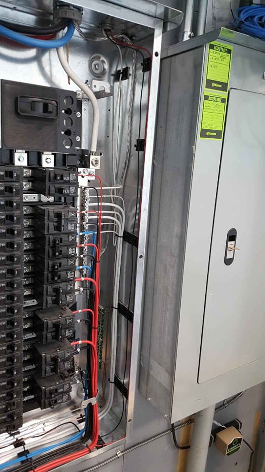 An open electrical service panel showing breakers, various colored wiring, and a closed adjacent metal subpanel.