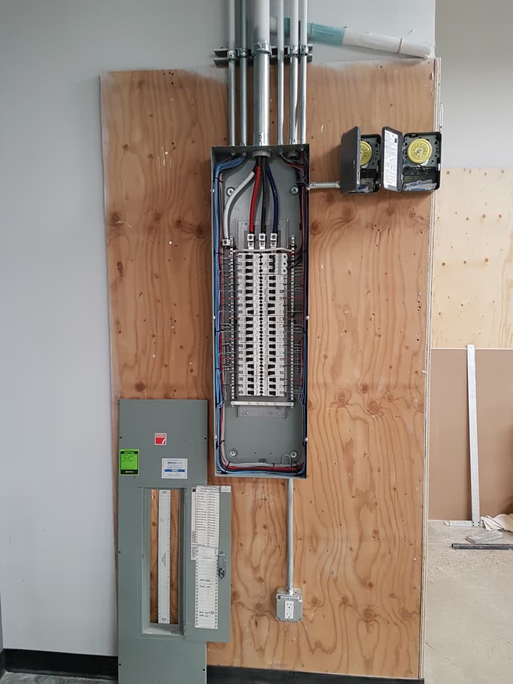 An electrical panel with the cover removed, mounted on a plywood wall with metal conduit and two small junction boxes.