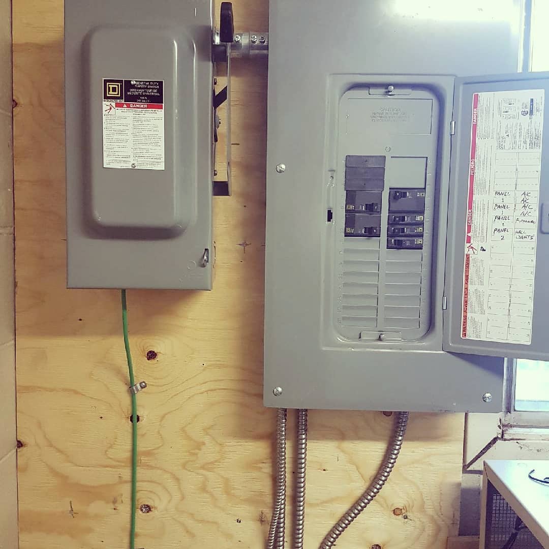 A gray electrical disconnect box and circuit breaker panel mounted on a plywood wall.