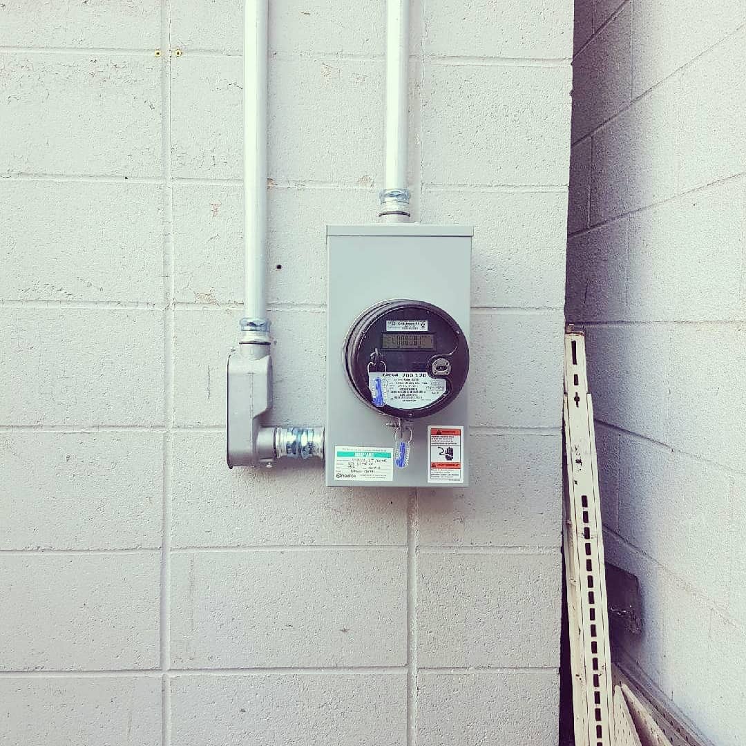 A gray electric utility meter mounted on a light-colored concrete block wall with two vertical metal conduits.
