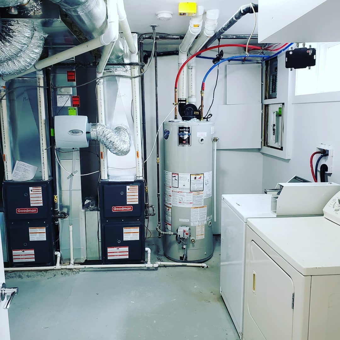 A basement utility room featuring two HVAC units, a water heater, and a washing machine against a white wall.