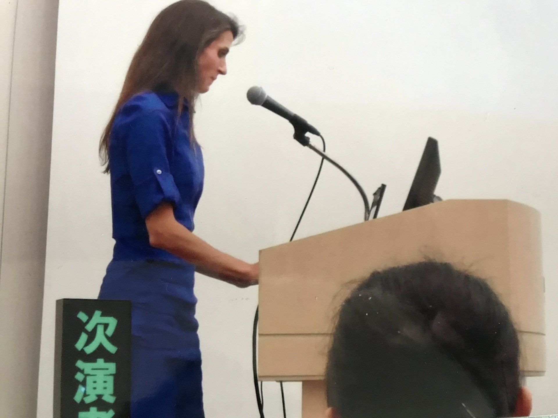 Woman speaking at a podium with a microphone, wearing a blue dress and looking at a laptop screen