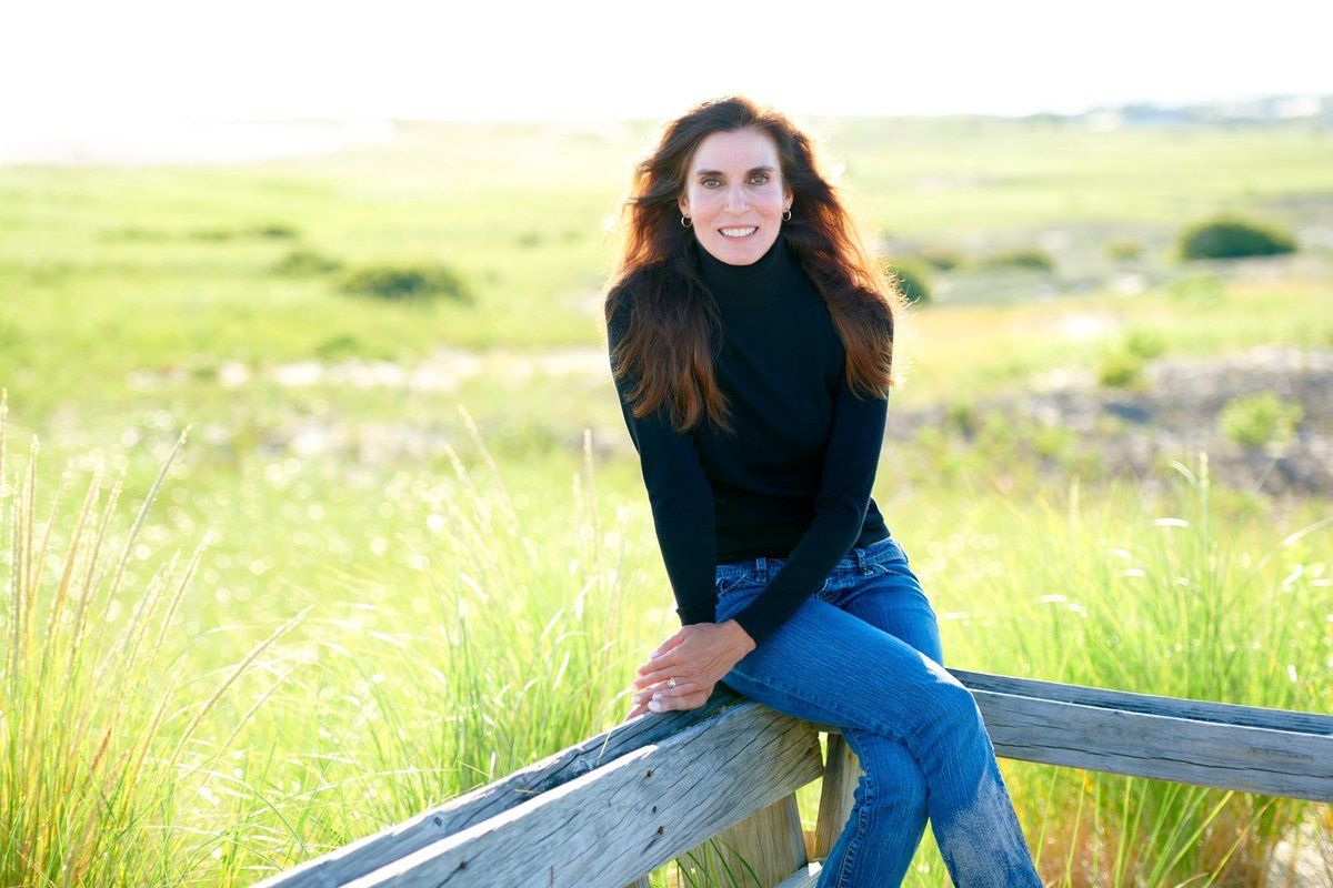 Woman in a black sweater and jeans sitting on a wooden fence in a sunny grassy field.