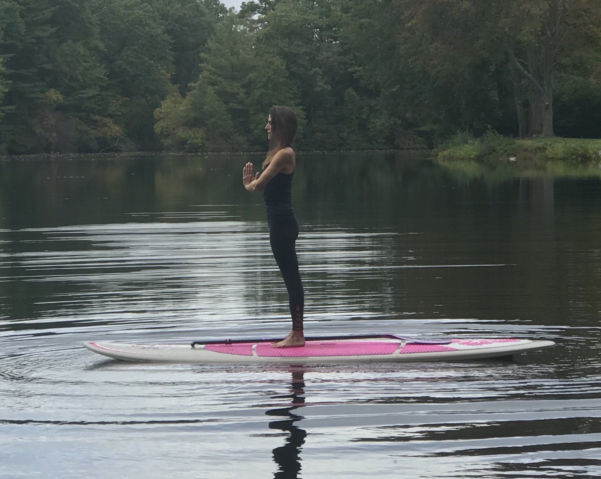 Person standing on a pink paddleboard on a calm lake, surrounded by trees.