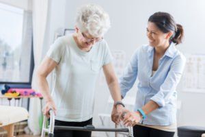 Caregiver helping an elderly person walk with a walker in a bright room
