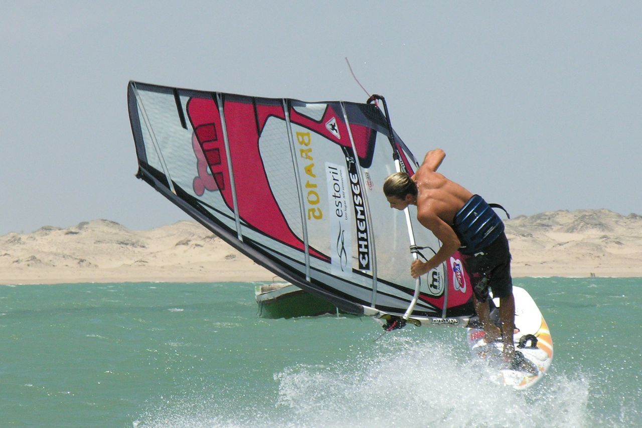 Um praticante de windsurf inclina-se para trás enquanto salta sobre as águas verdes e agitadas, segurando uma grande vela rosa e branca contra a costa arenosa.