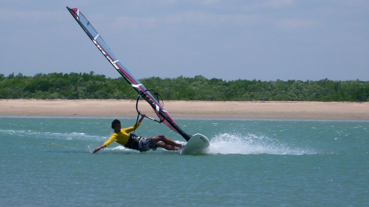 Uma pessoa com uma camisa amarela praticando windsurf em águas turquesas perto de uma praia de areia com árvores verdes ao fundo.