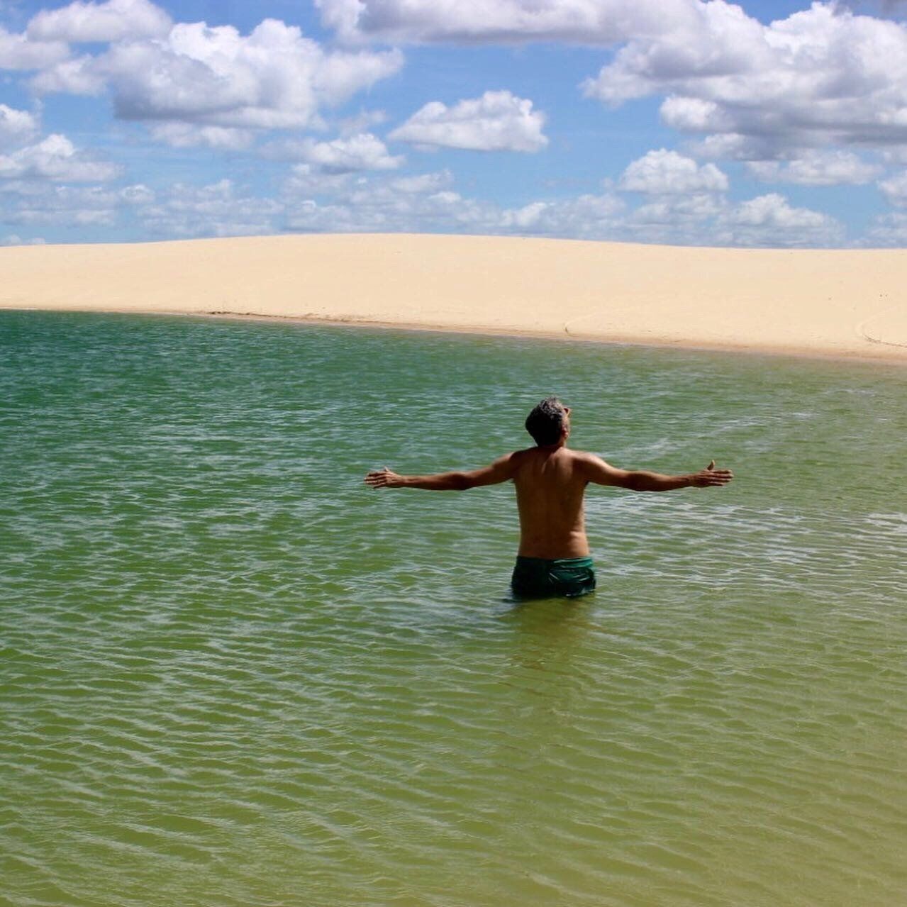 Uma pessoa está de pé em uma lagoa verde e cristalina, com os braços estendidos, de frente para uma grande duna de areia dourada sob um céu azul.