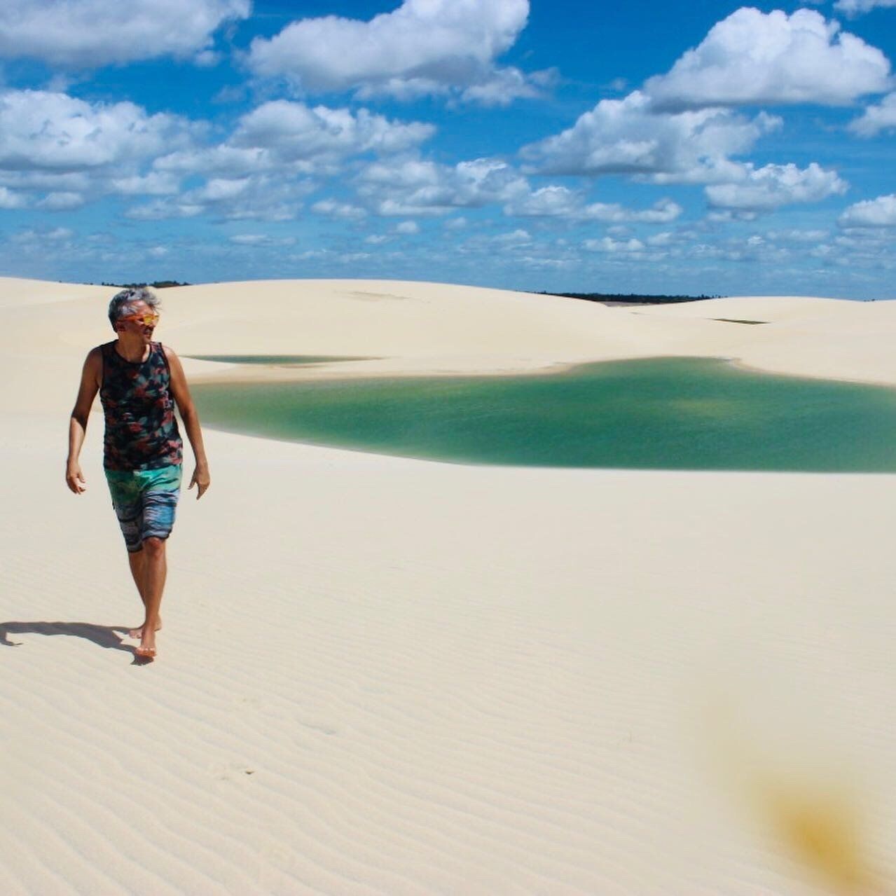 Uma pessoa caminha descalça pelas dunas de areia branca perto de uma lagoa turquesa sob um céu azul brilhante e nublado.