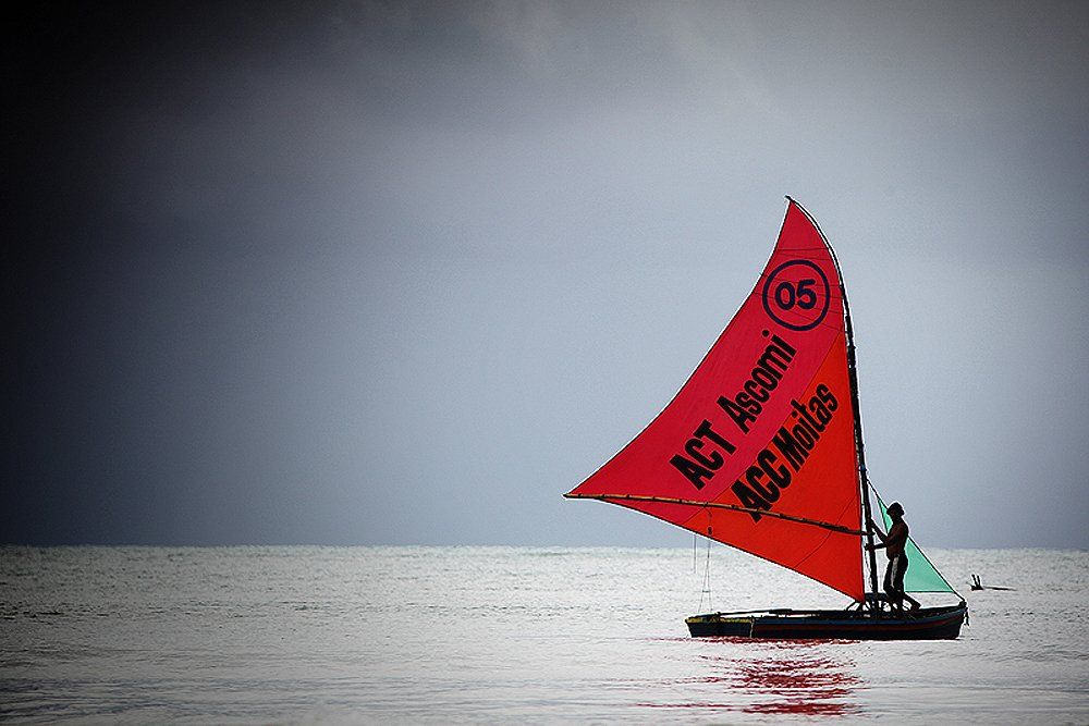 Uma vela vermelha com texto preto e o número 05 em um pequeno barco no oceano contra um céu escuro e tempestuoso.