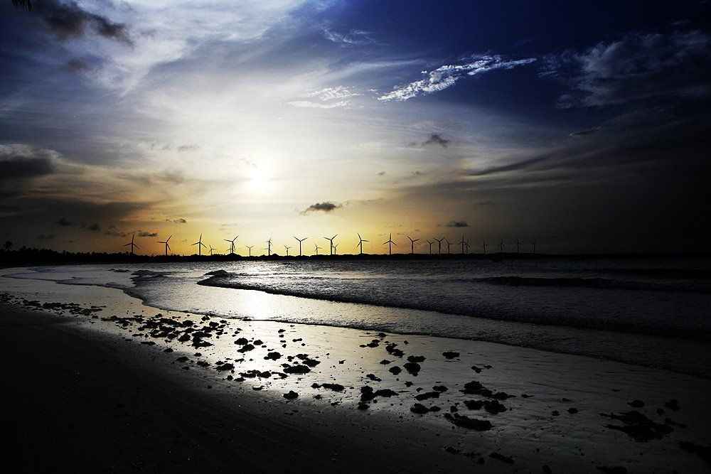 Ao pôr do sol, as silhuetas das turbinas eólicas se destacam na praia, com a areia escura e as ondas do oceano refletindo o céu claro.