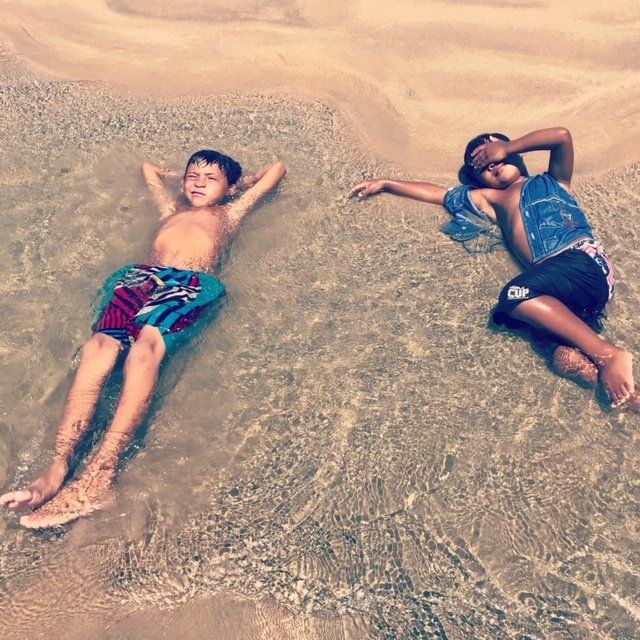 Duas pessoas estão deitadas de costas em águas rasas e cristalinas em uma praia de areia, relaxando com as mãos atrás da cabeça.