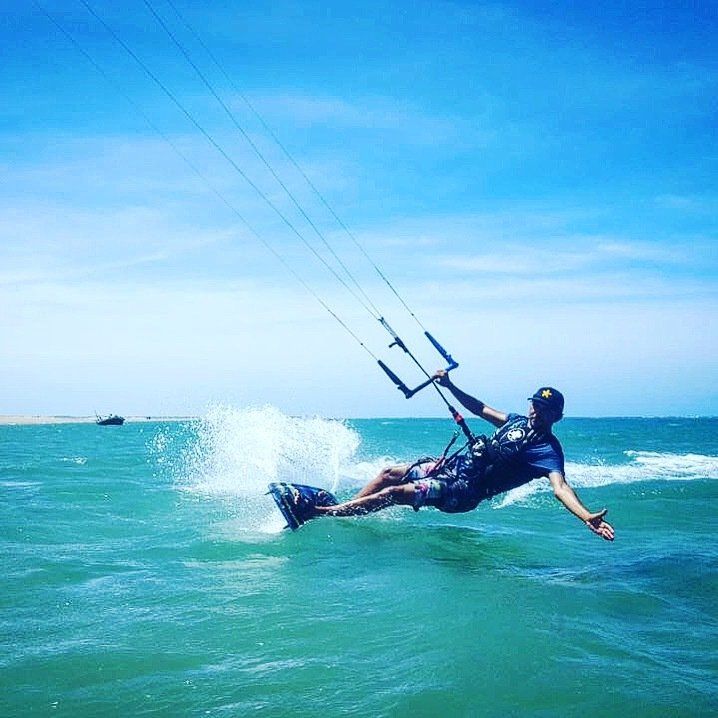 Uma pessoa praticando kitesurf em águas azuis e cristalinas, inclinada para trás com uma das mãos estendida enquanto segura a barra de controle.
