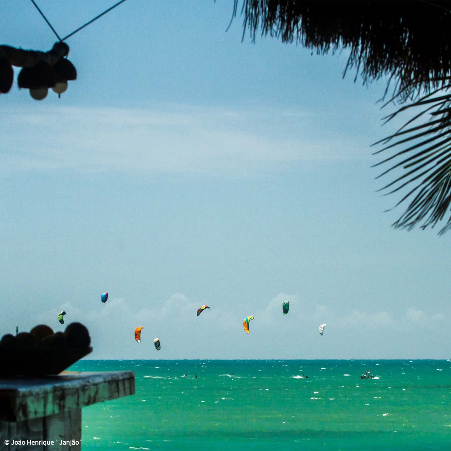 Inúmeras pipas coloridas voam sobre um oceano turquesa sob um céu claro, vistas de baixo de uma estrutura rústica e sombreada na praia.