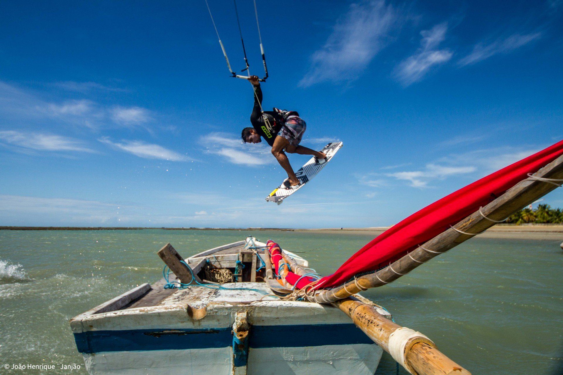 Um praticante de kitesurf salta sobre um pequeno barco de madeira com uma vela vermelha em águas calmas e ensolaradas sob um céu azul.