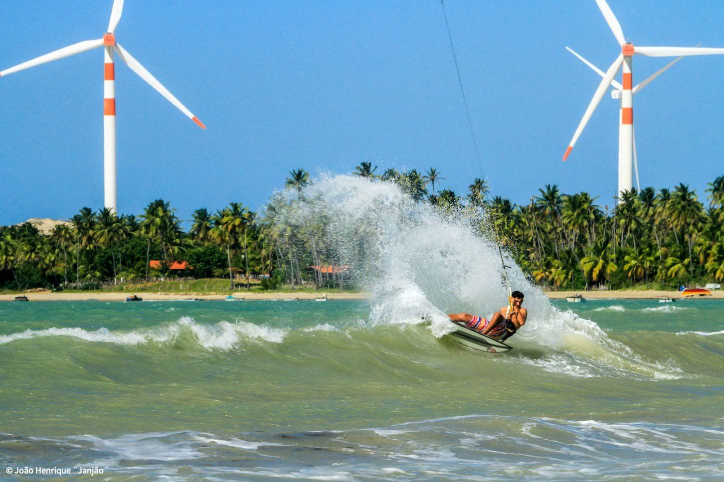 Uma pessoa praticando kitesurf em uma onda perto de uma costa tropical, com palmeiras e turbinas eólicas ao fundo.