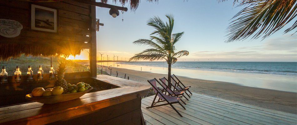 Um bar de praia de madeira ao pôr do sol, com espreguiçadeiras em um deck com vista para o oceano.
